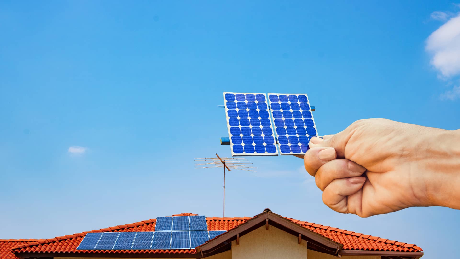 Mano gigante colocando un pequeño panel solar sobre el tejado de una casa representando un completo kit solar con baterías