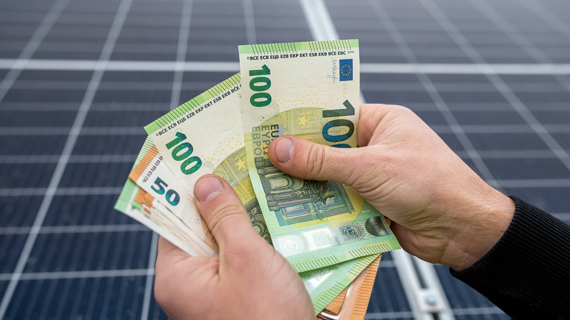 Mano sujetando billetes con placas solares de fondo ilustra el precio de la energía solar