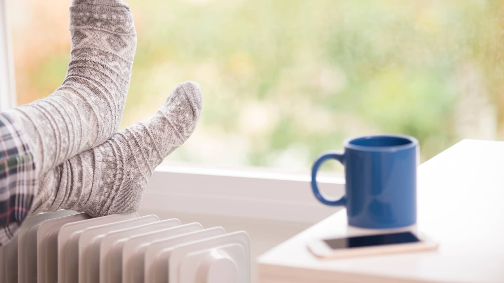 Persona descansa los pies en un radiador mientras su taza y móvil reposan en la mesilla, siguiendo nuestros consejos conseguira ahorrar en su calefacción
