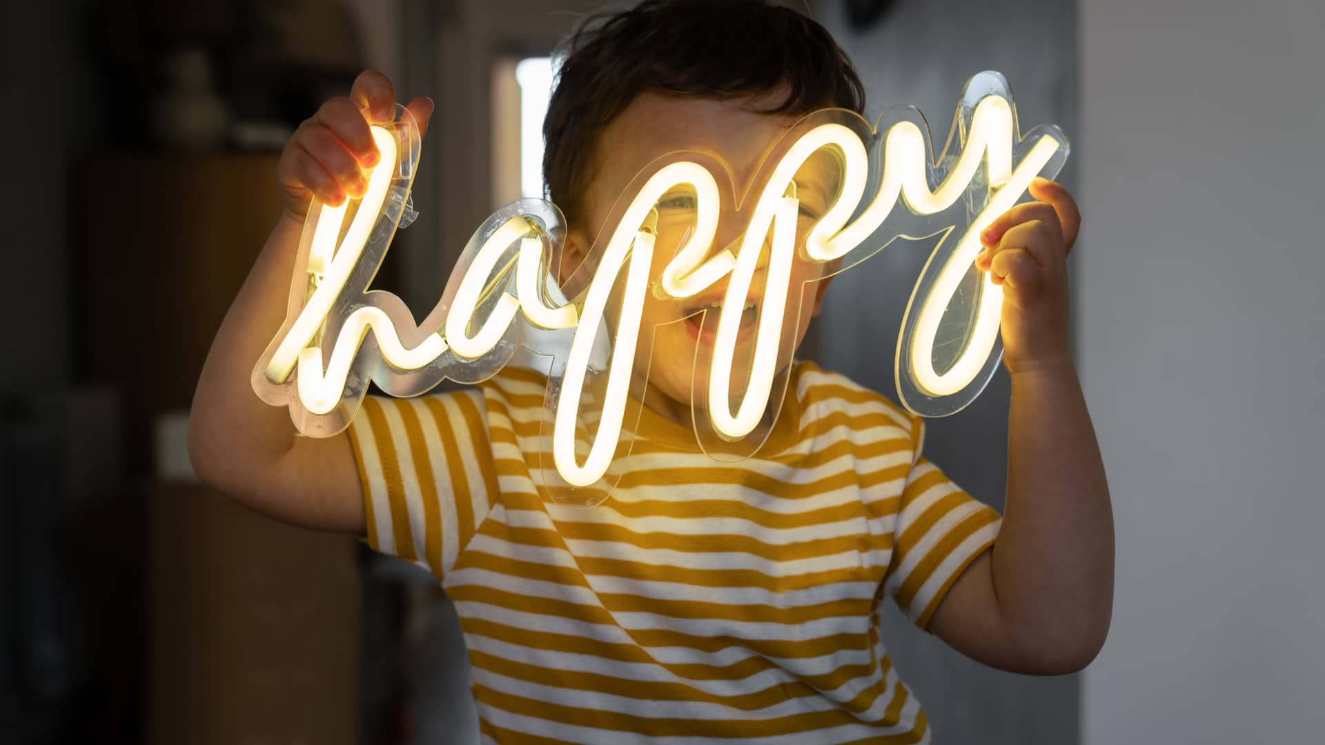 Niño sonriendo sosteniendo palabra luminosa LED happy, un tipo de luz que permite ahorrar sobre otras
