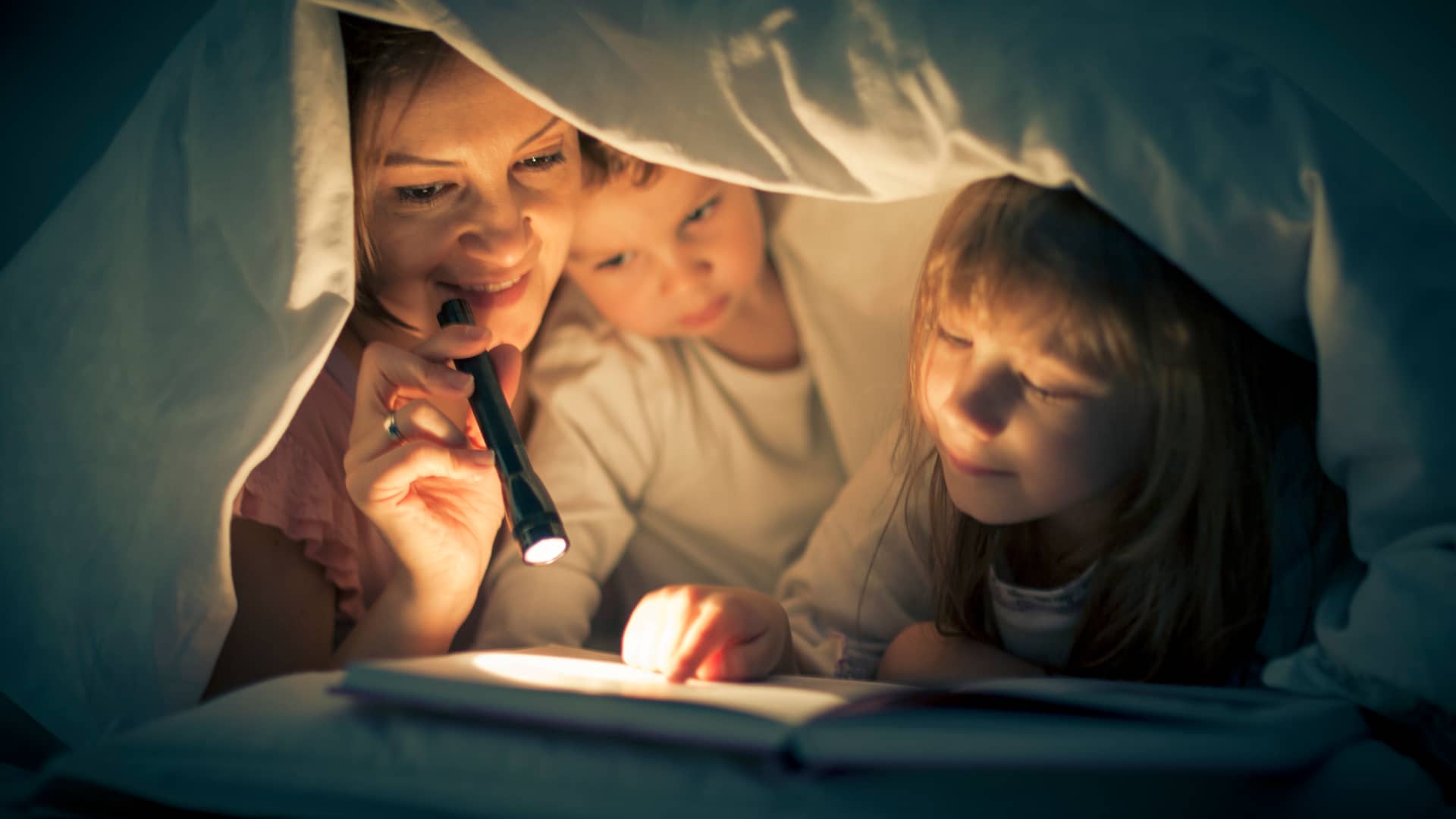 Madre leyendo un libro con sus hijos bajo la manta con la luz de una linterna debido al corte de luz
